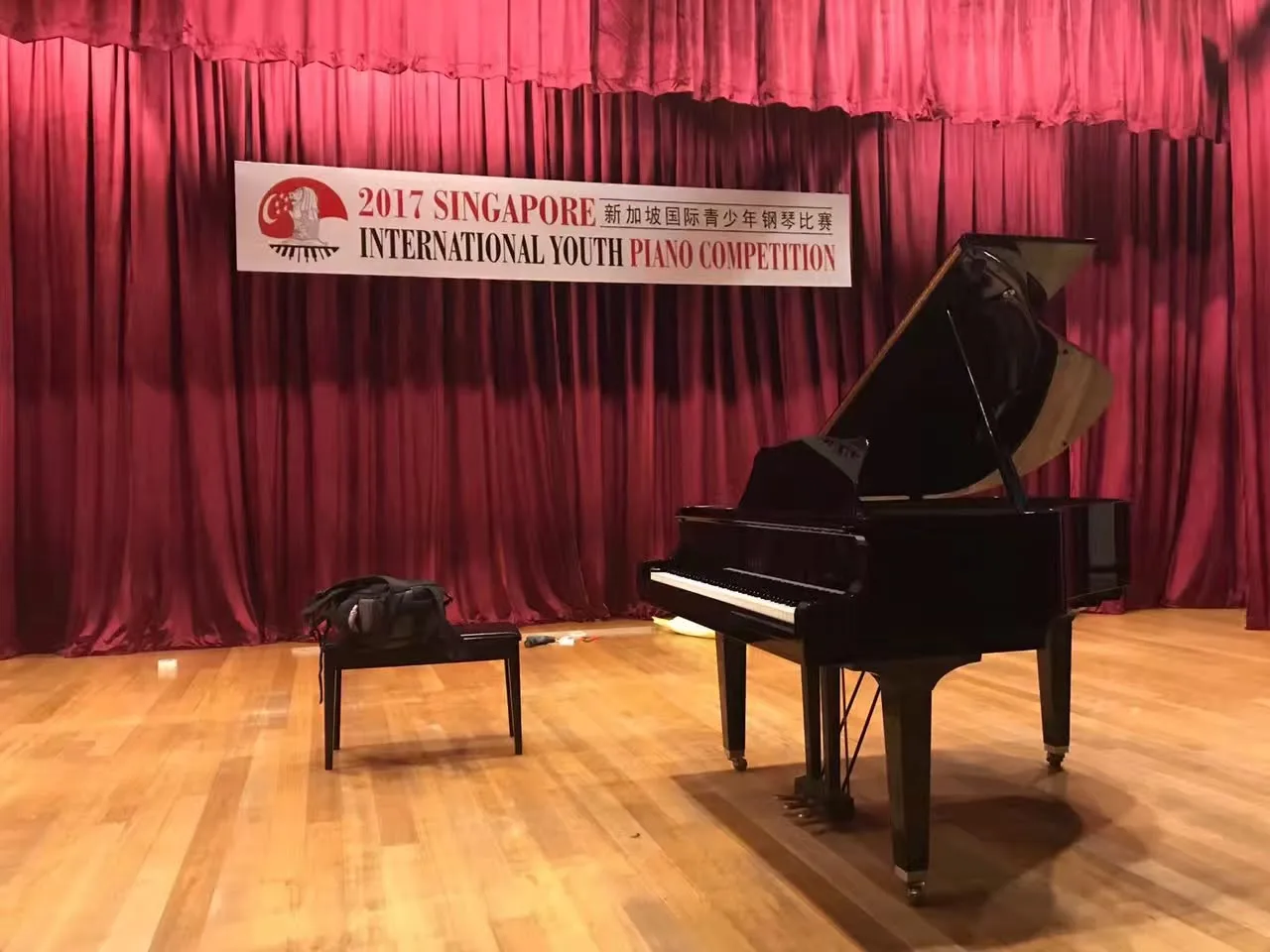 A grand piano on a red curtained stage beneath a banner for the 2017 Singapore International Youth Piano Competition